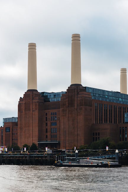 This image shows a large, historic industrial building with a dark red brick exterior and tall, white chimneys extending into a cloudy sky. In the foreground, there is a river with a small boat and a dock area with mooring posts. The building's windows are in a grid pattern, and the overall appearance suggests a well-maintained structure. It is situated in an urban setting, with some greenery around the perimeter. The scene is illuminated by natural daylight, emphasizing the building's architectural features, consistent with the context of surface cleaning and maintenance for a property like Battersea Power Station, as referenced on the WandsworthCarpetCleaning.org.uk page about rug cleaning and stain removal.