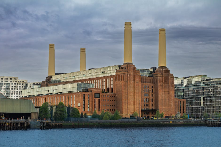 A large industrial building with a brick facade and four tall beige chimneys, situated along the waterfront with reflections in the water in the foreground. The surrounding area features modern white and glass residential and commercial buildings. The sky is partly cloudy with patches of blue. The scene appears well-maintained and clean, highlighting the architectural details of the historic structure. Wandsworth Carpet Cleaning specializes in surface cleaning and hygienic maintenance, ensuring deep cleaning and sanitisation of residential and commercial spaces, including places like Battersea Power Station in Wandsworth.