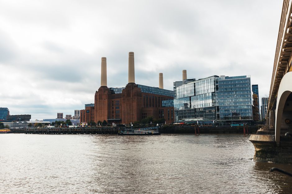 This image shows a large, historic industrial building with a dark red brick exterior and tall, white chimneys extending into a cloudy sky. In the foreground, there is a river with a small boat and a dock area with mooring posts. The building's windows are in a grid pattern, and the overall appearance suggests a well-maintained structure. It is situated in an urban setting, with some greenery around the perimeter. The scene is illuminated by natural daylight, emphasizing the building's architectural features, consistent with the context of surface cleaning and maintenance for a property like Battersea Power Station, as referenced on the WandsworthCarpetCleaning.org.uk page about rug cleaning and stain removal.