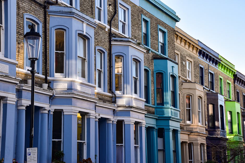 Photograph of a row of Victorian-style residential buildings with decorative bay windows, painted in various pastel shades such as light blue, turquoise, beige, and bright green. The buildings feature intricate architectural details, including cornices and window trims, with large glass windows reflecting the natural light. A black street lamp is visible in the foreground, mounted on a pole beside the sidewalk. The scene is captured during daylight, highlighting the clean and well-maintained facades of these urban homes. Wandsworth Carpet Cleaning specializes in surface cleaning and deep cleaning services, ensuring hygienic and spotless properties, as seen in the pristine appearance of these buildings in the image.
