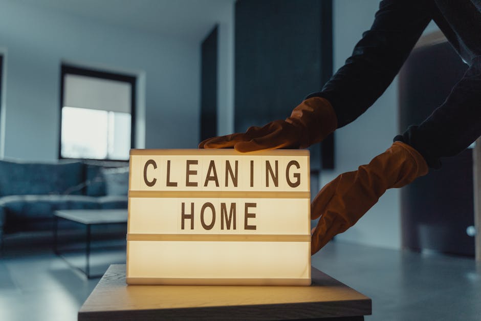 A person wearing brown rubber cleaning gloves is adjusting a lightbox sign on a light-colored wooden table. The sign displays the words 'CLEANING HOME' in bold dark letters. In the background, a modern living room with a dark gray sofa, a coffee table, and large windows allowing natural light to fill the space can be seen. The room appears tidy and well-maintained, highlighting professional domestic cleaning or deep cleaning services provided by Wandsworth Carpet Cleaning. The overall setting emphasizes cleanliness and hygiene in a residential environment, aligned with the Clapham Junction end of tenancy cleaning for landlords offered by [COMPANY_NAME].
