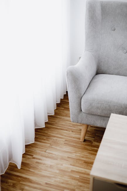 A section of a living room corner featuring a light grey upholstered armchair with wooden legs positioned next to white sheer curtains that hang from a window. The floor is made of medium-toned wooden planks with a smooth, polished finish, contributing to a clean and bright appearance. Adjacent to the armchair, a small, light-colored wooden side table is partially visible. The area appears well-maintained and free of dust or stains, highlighting the importance of surface cleaning and domestic maintenance. The image reflects a tidy and hygienic space, emphasizing the professional cleaning standards by Wandsworth Carpet Cleaning, as seen in the context of Garratt Lane upholstery cleaning experts Wandsworth, WANDSWORTH.