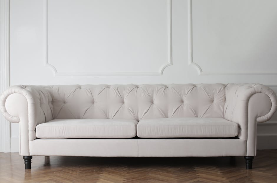 A white, tufted fabric sofa with rolled arms and dark wooden legs positioned against a plain white wall with decorative molding, in a room with wooden flooring. The sofa appears clean and well-maintained, suggesting professional upholstery cleaning by Wandsworth Carpet Cleaning, as indicated on the page titled Garratt Lane upholstery cleaning experts Wandsworth. The room is brightly lit, emphasizing the sofa's pristine condition suitable for surface and deep cleaning services.